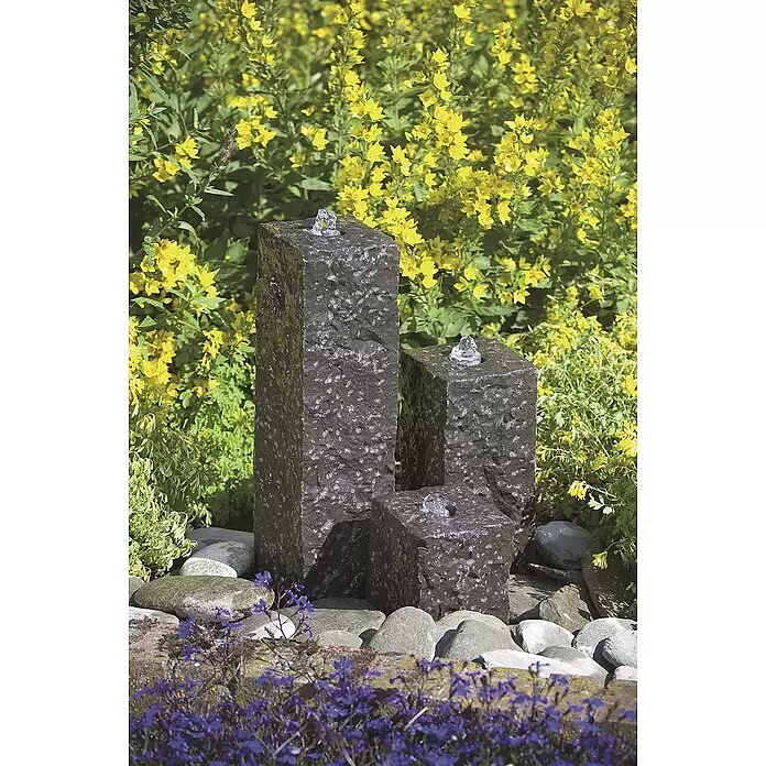 Ubbink AcquaArte Gartenbrunnen Modena - 12 x 12 x 45 cm, Naturmaterialien, Anthrazit - Drei dunkle, strukturierte Steinsäulenbrunnen unterschiedlicher Höhe, mit sprudelndem Wasser, zwischen Felsen und Gartenblumen.