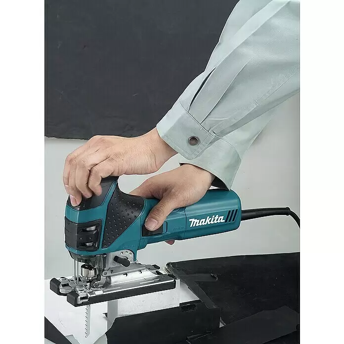 Makita Caladora eléctrica 4351FCTJ - 800 /min - 2.800 /min, Profundidad de corte máx. en madera: 135 mm, Número de niveles de elevación del péndulo: 4 ud. - Sierra de calar Makita turquesa y negra sostenida por manos, cortando un material negro.
