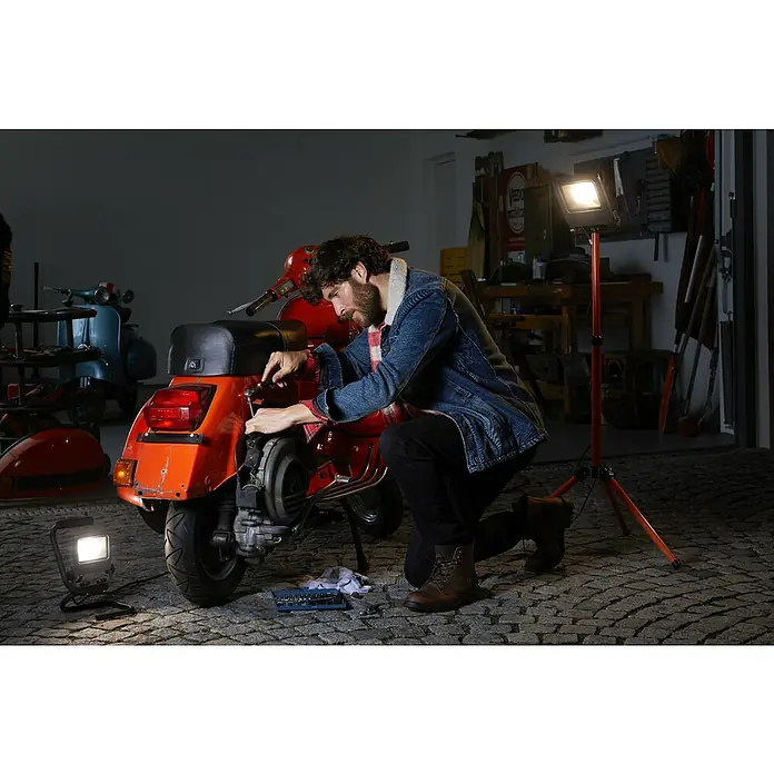 Ledvance LED-Arbeitsleuchte - 1.700 lm, Lichtfarbe: Kaltweiß, Anzahl Leuchtmittel: 1 Stk. - Ein Mann repariert einen orangefarbenen Motorroller in einer Garage, beleuchtet von Arbeitsleuchten, Werkzeug liegt daneben.