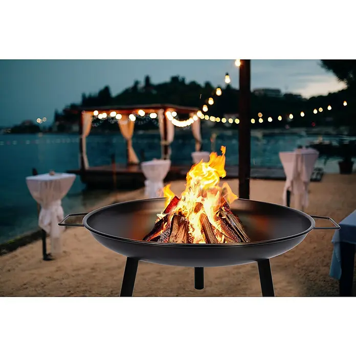 Brasero Fulton - Diámetro: 60 cm, Acero - Un brasero con leña encendida en una zona de arena, con un bar de playa iluminado y mesas al fondo.