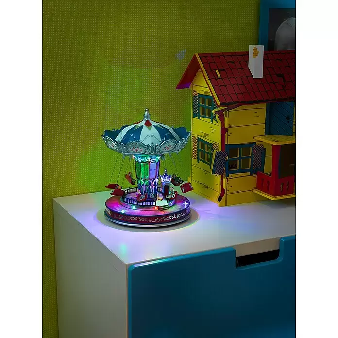 Konstsmide LED-Szenerie Karussell bunt - L x B x H: 21 x 21 x 26,5 cm, Bunt, Netzbetrieben, LED - Ein buntes, beleuchtetes Karussellspielzeug mit Schaukeln neben einem gelben Spielzeughaus mit rotem Dach auf einer weißen Oberfläche.