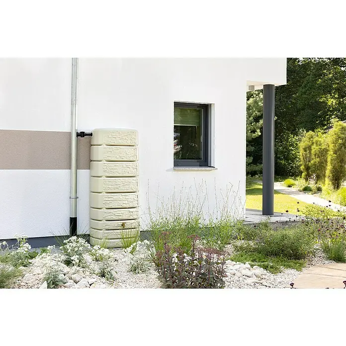 4rain Regenspeicher Slim Stone - 500 l, Beige - Ein stilvoller, beigefarbener Wasserspeicher steht neben einem Regenrohr an einer Hauswand, umgeben von üppigem Grün und weißen Steinen.