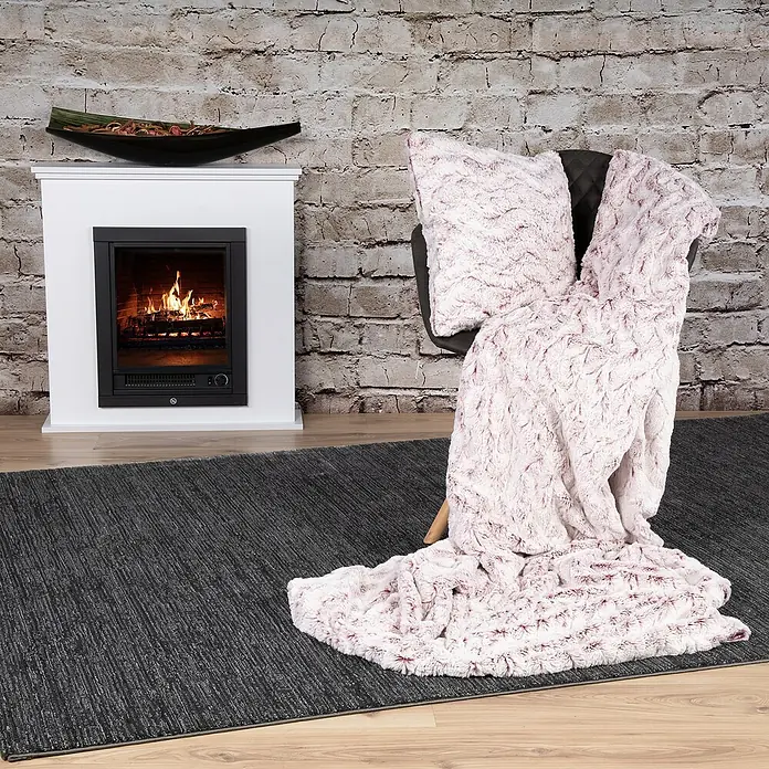 Decke Smooth - Rot, 200 x 150 cm, 100 % Polyester - Ein weicher, rosafarbener Überwurf und ein Kissen liegen auf einem Stuhl neben einem modernen Kamin, der eine gemütliche Atmosphäre schafft.
