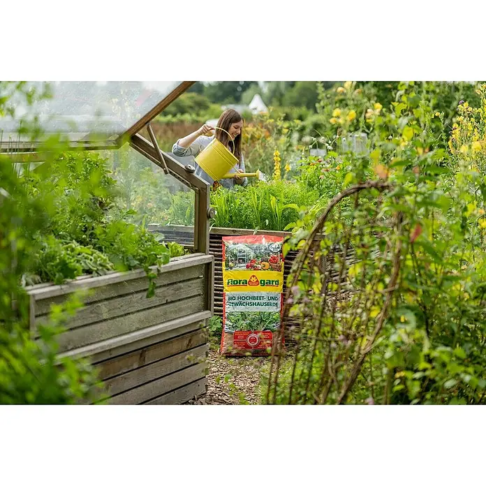 Floragard Bio-Hochbeeterde - 70 l - Flora Gard BIO Hochbeet- und Gewächshauserde neben einem Hochbeet und einer Frau, die mit einer gelben Gießkanne gießt.