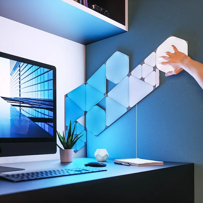 Nanoleaf Erweiterungskit Shapes Hexagons 3 Panels - 3 Stk., Weiß, RGBW, Hexagons, Smart Home-fähig: Ja - Intelligente LED-Wandpaneele in geometrischen Formen an einer blauen Wand über einem Schreibtisch.