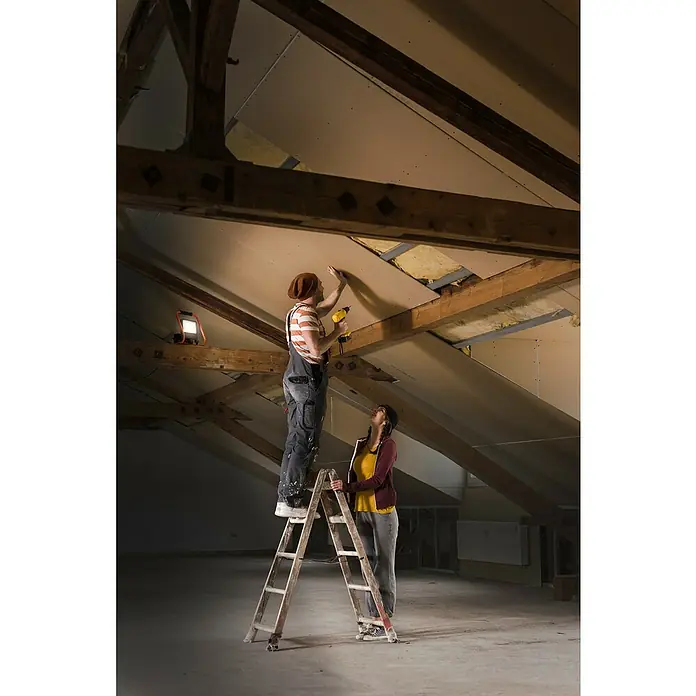 Ledvance LED-Strahler - 30 W, 2.700 lm, 4.000 K, L x B x H: 19 x 22 x 29 cm, IP44 - Mann befestigt Gipskartonplatten mit einem gelben Akku-Bohrschrauber an einer Dachschräge, während eine Frau die Leiter hält.