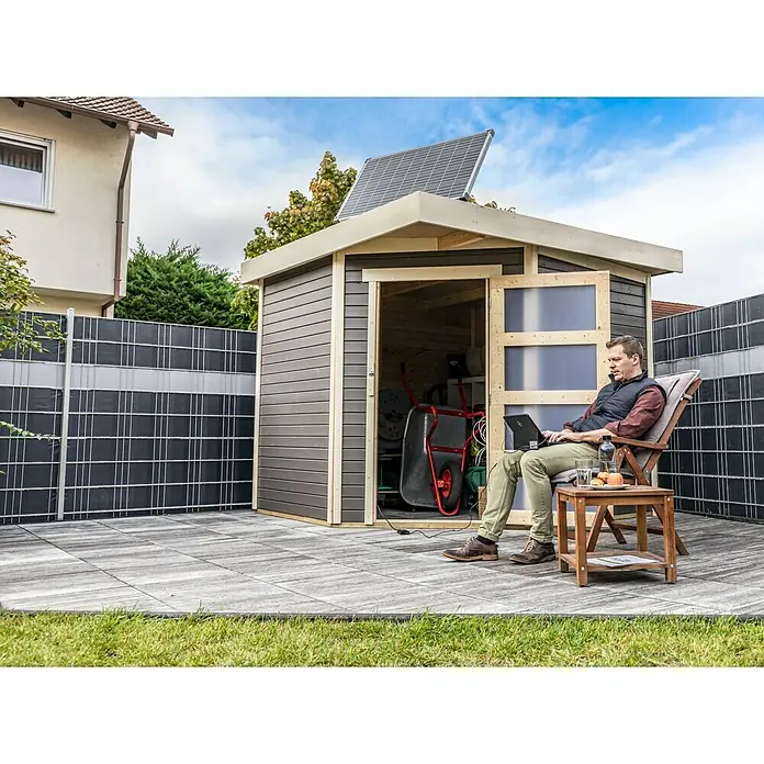Offgridtec Solarbatterie - 122 Ah, 12 V - Ein modernes Gartenhaus mit Solarmodul auf dem Dach, neben einem Mann, der entspannt mit seinem Laptop arbeitet.