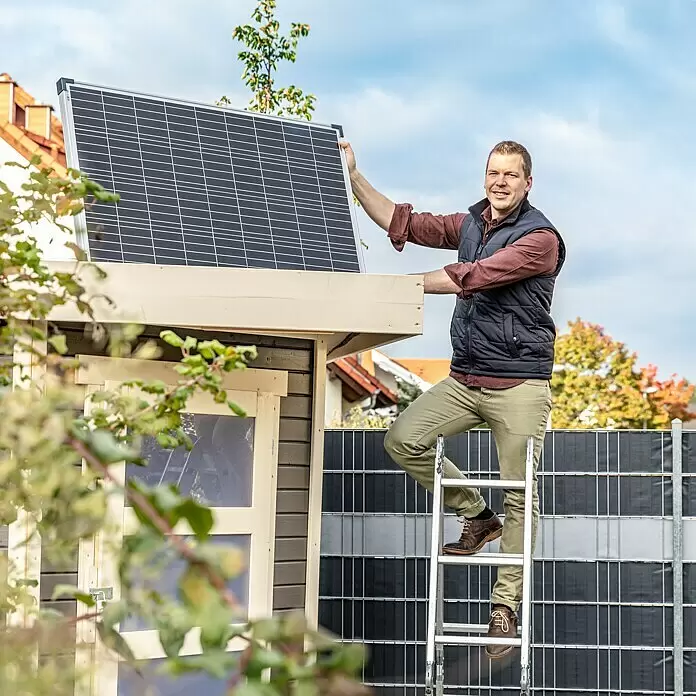 Offgridtec Solarmodul 0% MwSt. (gem. § 12 Abs. 3 UStG) - Nennleistung: 150 W, 114 x 68 x 3,5 cm, 1 Stk. - Mann montiert ein dunkles Solarmodul mit silbernem Rahmen auf einem Schuppendach.