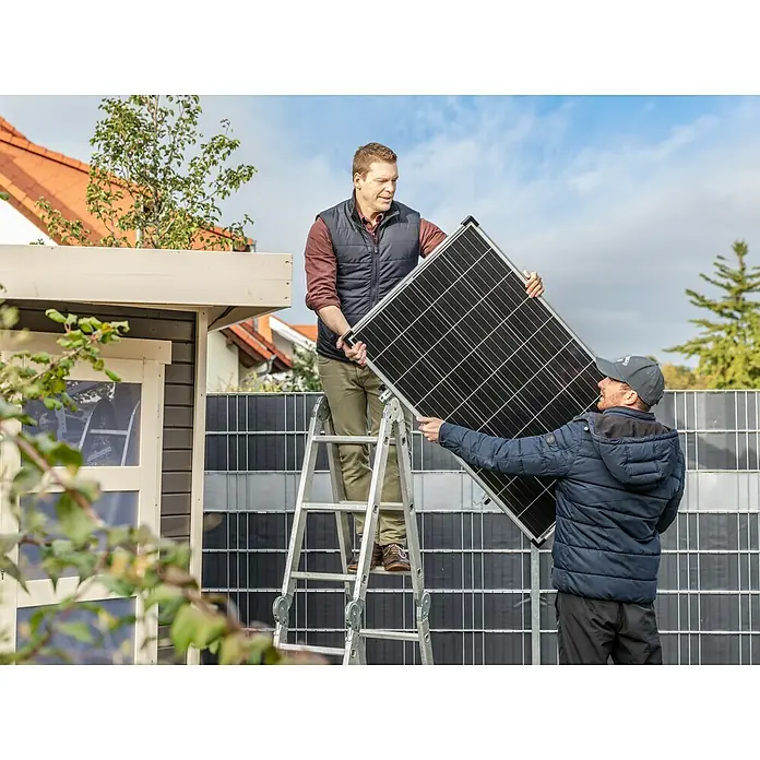 Offgridtec Solarmodul 0% MwSt. (gem. § 12 Abs. 3 UStG) - Nennleistung: 150 W, 114 x 68 x 3,5 cm, 1 Stk. - Zwei Männer installieren ein Solarmodul auf einem Dach, wobei einer auf einer Leiter steht und der andere es hält.