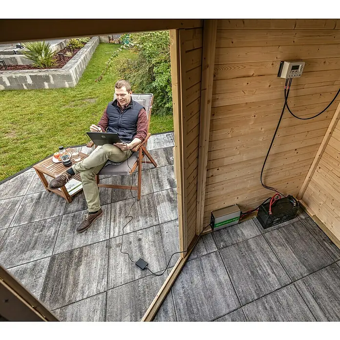 Offgridtec Batteriekabel Verbindungskabel - Länge: 40 cm, Querschnitt: 25 mm², Schwarz - Ein Mann sitzt im Garten vor einer Holzhütte mit Solaranlage, bestehend aus Laderegler, Batterie und Wechselrichter, und arbeitet am Laptop.