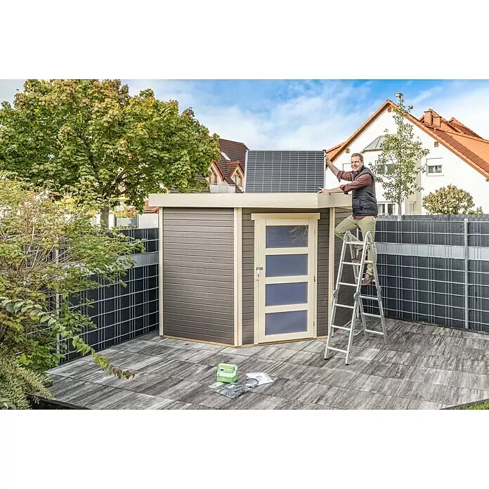 Offgridtec Solarmodul 0% MwSt. (gem. § 12 Abs. 3 UStG) - Nennleistung: 150 W, 114 x 68 x 3,5 cm, 1 Stk. - Mann installiert schwarzes Solarpanel auf grauem Gartenhaus mit heller Verkleidung und Tür in einem gepflasterten Garten.