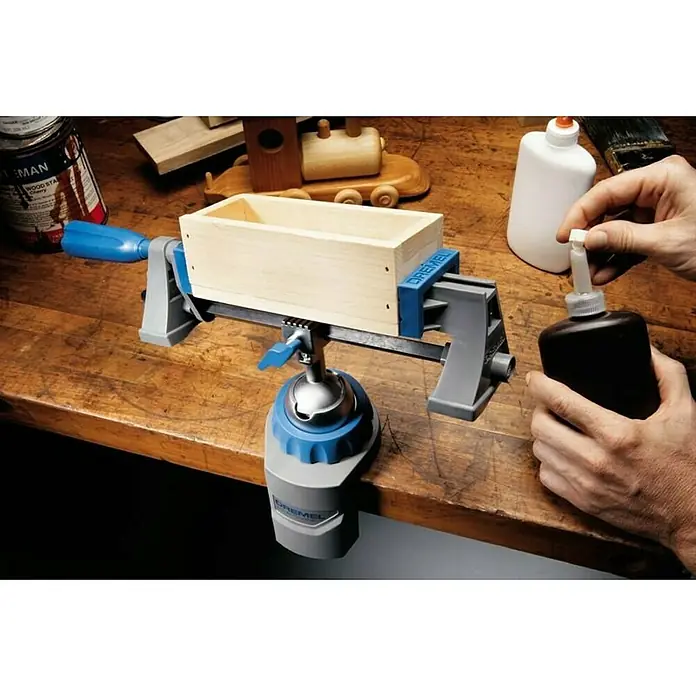 Dremel Multi-Schraubstock 2500 - Neigungswinkel 50 ° - Ein Dremel Werkstückhalter hält eine Holzkiste, während eine Person eine Flasche Kleber hält.