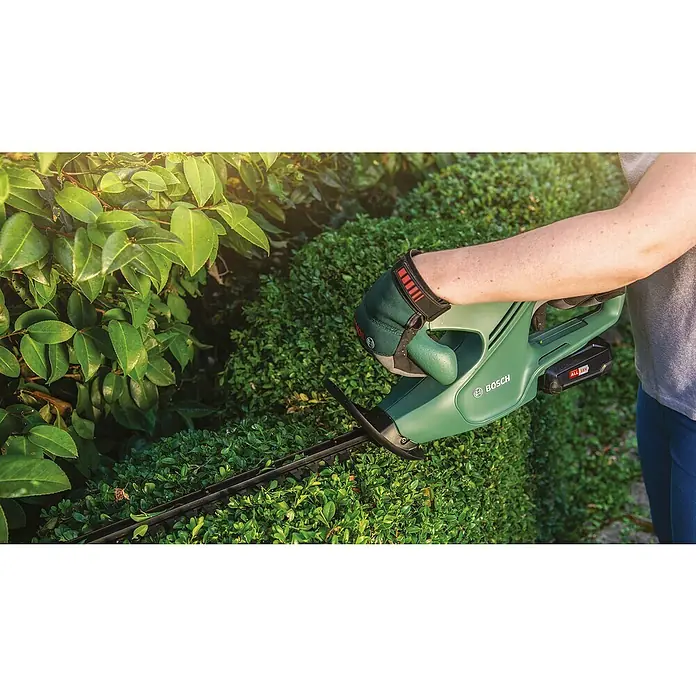 Bosch Power for All 18V Akku-Heckenscheren-Set EasyHedgeCut 18-45 + ALB 18 LI - 18 V, 2 Ah, Li-Ionen - Eine grüne Bosch-Akku-Heckenschere schneidet eine Hecke, wobei die Hand der Person einen Handschuh trägt.
