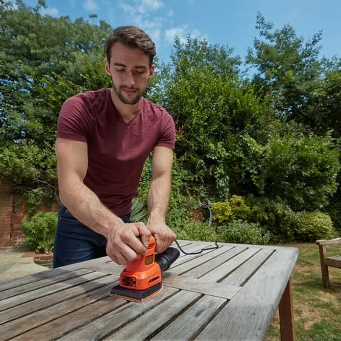 Black+Decker Lijadora multifunción eléctrica  BEW200LSA + 21 accesorios y bolsa - 200 W, Número de oscilaciones marcha en vacío: 12.000 r.p.m., Diámetro circuito oscilante: 2 mm - Hombre lijando una superficie de madera al aire libre con una lijadora Black+Decker.