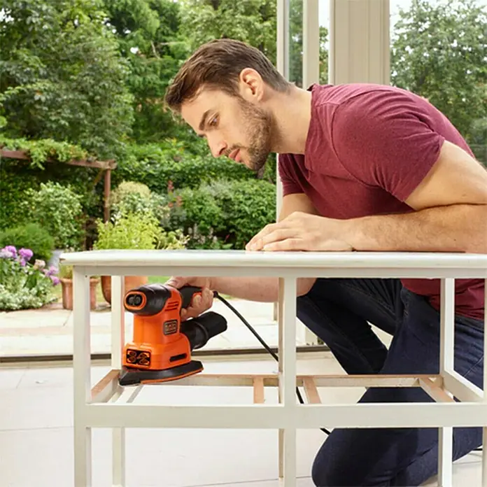 Black+Decker Lijadora multifunción eléctrica  BEW200LSA + 21 accesorios y bolsa - 200 W, Número de oscilaciones marcha en vacío: 12.000 r.p.m., Diámetro circuito oscilante: 2 mm - Un hombre lija una mesa con una lijadora eléctrica Black+Decker. La lijadora es naranja y negra.
