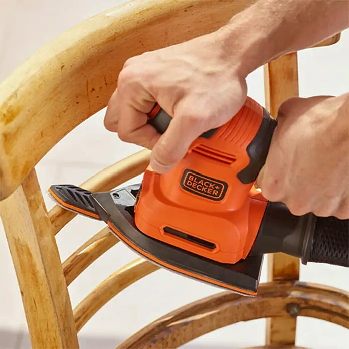 Black+Decker Lijadora multifunción eléctrica  BEW200LSA + 21 accesorios y bolsa - 200 W, Número de oscilaciones marcha en vacío: 12.000 r.p.m., Diámetro circuito oscilante: 2 mm - Primer plano de un hombre lijando una silla de madera con una lijadora de detalles naranja Black+Decker.