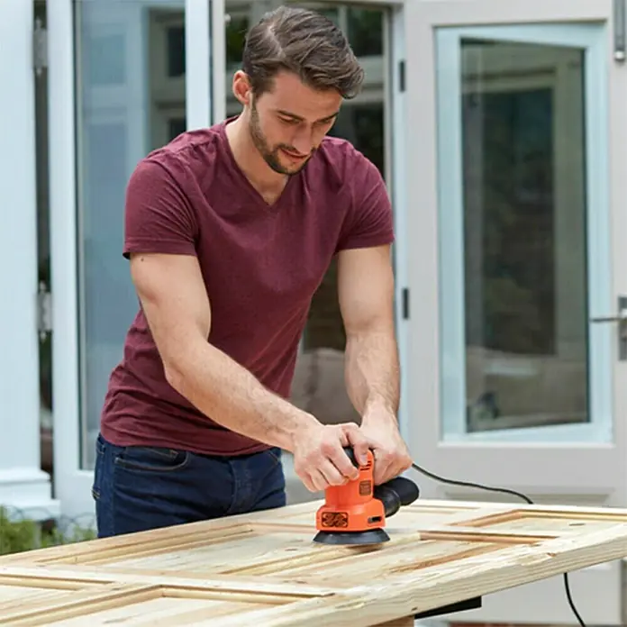 Black+Decker Lijadora multifunción eléctrica  BEW200LSA + 21 accesorios y bolsa - 200 W, Número de oscilaciones marcha en vacío: 12.000 r.p.m., Diámetro circuito oscilante: 2 mm - Un hombre utiliza una lijadora orbital Black+Decker para trabajar en una puerta de madera al aire libre. La herramienta es naranja y negra.