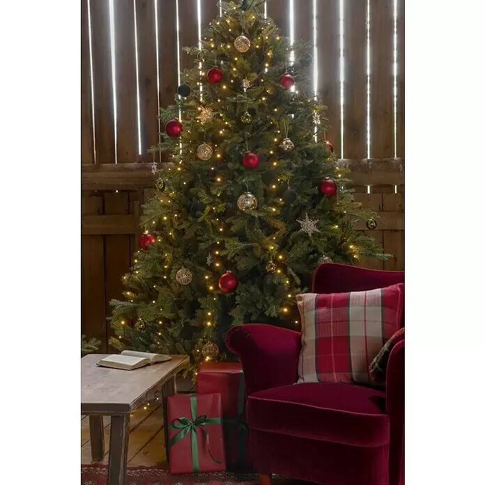 Konstsmide Baummantel - Innen, 2,4 m, 5 Stränge à 40 gefrostete bernsteinfarbene LEDs - Ein festlicher Weihnachtsbaum, geschmückt mit Lichtern und Kugeln, steht neben einem roten Samtsessel mit Plaidkissen und Geschenken.