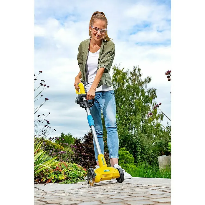Gloria Power for All 18V Fugenreiniger WeedBrush li-on - 18 V, Li-Ionen, Ohne Akku - Frau benutzt einen gelben Gloria Steinreiniger auf einem Gehweg, um Unkraut zu entfernen. Ideal für Gartenarbeiten und die Pflege von Außenbereichen.