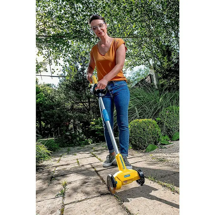 Gloria Power for All 18V Fugenreiniger WeedBrush li-on - 18 V, Li-Ionen, Ohne Akku - Frau benutzt einen Gloria Multibrush Kehrbesen, um Unkraut von einer Terrasse zu entfernen. Das Gerät ist gelb und silber.