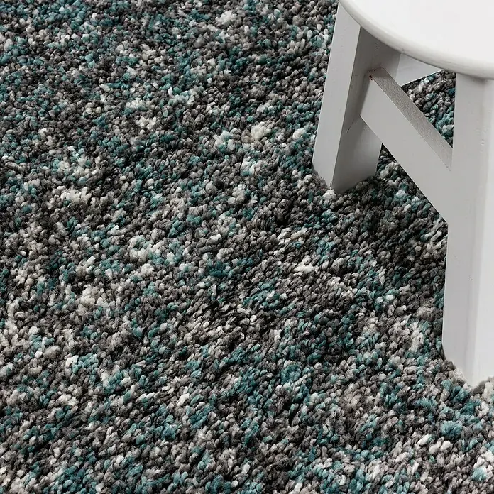 Hochflorteppich Enjoy 4500Blau, 290 x 200 cm, 100 % Polypropylen - Detailansicht eines mehrfarbigen Shaggy-Teppichs mit einem weißen Hockerbein. Der Teppich hat blaue, graue und weiße Farbtöne.