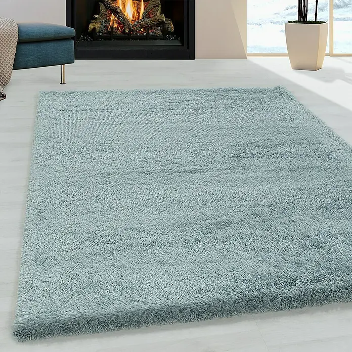 Hochflorteppich Fluffy 3500Blau, 340 x 240 cm, 100% Polypropylen - Ein quadratischer, hellblauer Shaggy-Teppich liegt in einem hellen Raum mit Kamin und Sitzmöbeln. Der Teppich hat eine weiche, flauschige Textur.