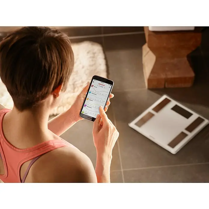 Báscula digital Shape Sense Connect 100 - 32 x 32 x 2,6 cm, Vidrio, Blanco - Mujer comprobando su peso y datos corporales en un teléfono inteligente, obtenidos de una báscula de análisis corporal Soehnle.