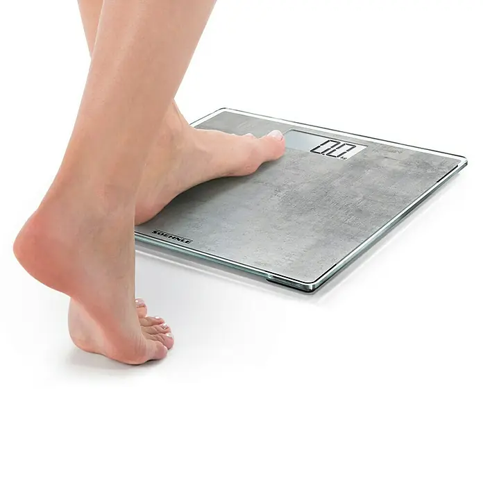 Báscula digital Style Sense Compact 300 - 29 x 29 x 2 cm, Vidrio, Gris hormigón - Persona de pie en una báscula Soehnle, con una superficie gris texturizada y una pantalla digital que muestra "0.0 kg".