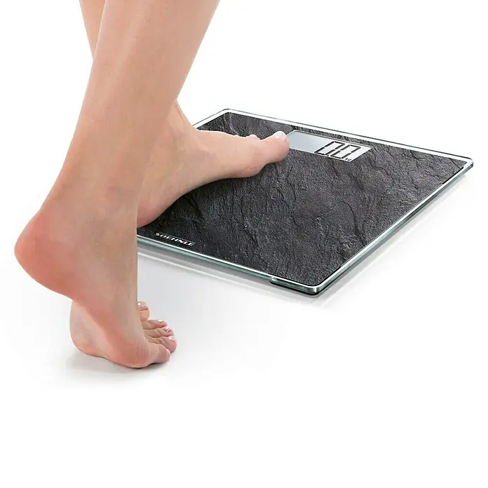 Báscula digital Style Sense Compact 300 - 29 x 29 x 2 cm, Vidrio, Negro pizarra - Pie sobre una báscula de baño digital negra con aspecto de piedra y borde de cristal, mostrando '0.0 kg'.