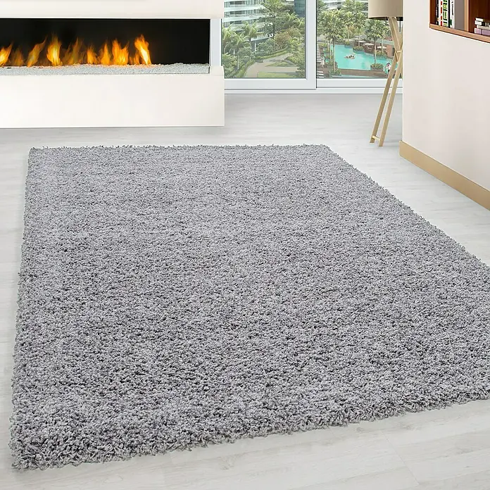 Hochflorteppich Life 1500 - Light Grey, 110 x 60 cm, 100 % Polypropylen - Ein grauer, weicher Shaggy-Teppich liegt in einem hellen Raum mit Kamin und großem Fenster mit Poolblick.