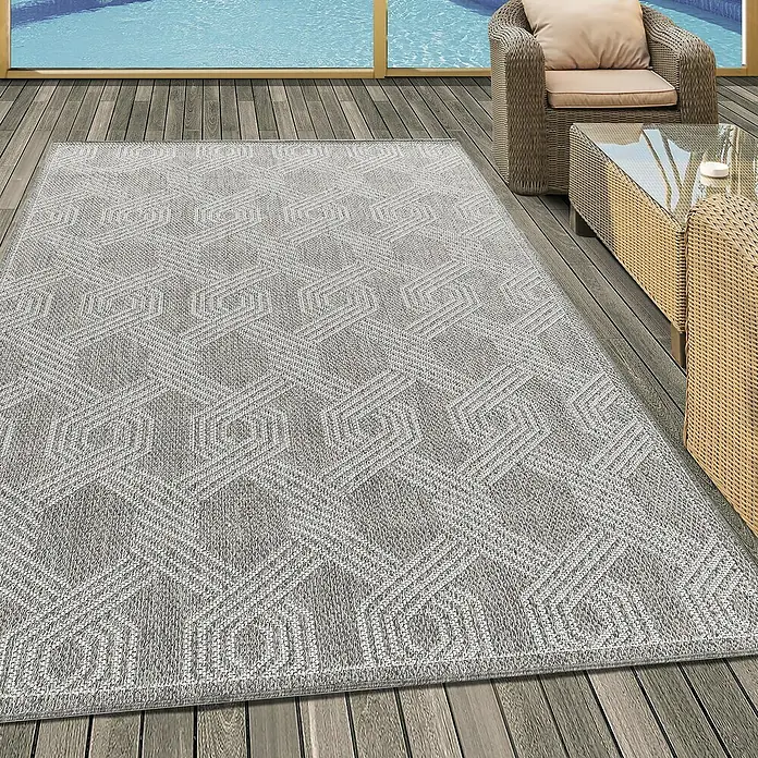 Outdoor-Teppich Aruba 4904Grau, 340 x 240 cm, 100% Polypropylen - Ein grauer Teppich mit geometrischem Muster liegt auf einer Holzterrasse, neben einem Rattansessel und einem Tisch mit Glasplatte.
