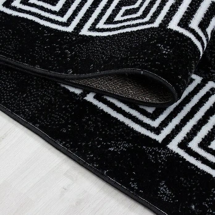 Kurzflorteppich Plus 8009 - Schwarz, 370 x 280 cm, 100 % Polypropylen - Nahaufnahme eines schwarzen Teppichs mit weißem geometrischem Muster und aufgerolltem Rand.