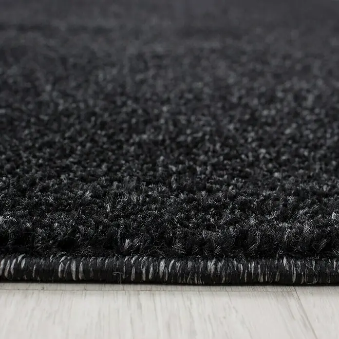 Kurzflorteppich Ata 7000Anthrazit, Durchmesser: 120 cm, 100 % Polypropylen - Nahaufnahme eines schwarzen Teppichs mit leichter Struktur, der auf einem hellen Holzboden liegt. Die Bordüre des Teppichs ist sauber verarbeitet.