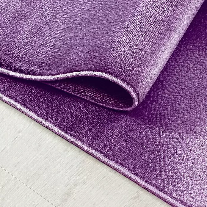 Kurzflorteppich Plus 8008 - Lila, 290 x 200 cm, 100% Polypropylen - Ein texturierter violetter Teppich, teilweise auf einem hellen Holzboden aufgerollt, zeigt seinen eingefassten Rand.