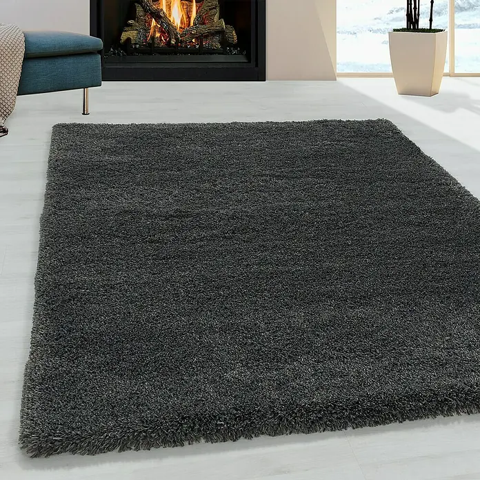 Hochflorteppich Fluffy 3500 - Grau, 170 x 120 cm, 100 % Polypropylen - Ein grauer, rechteckiger Hochflorteppich liegt auf einem hellen Holzboden vor einem Kamin.