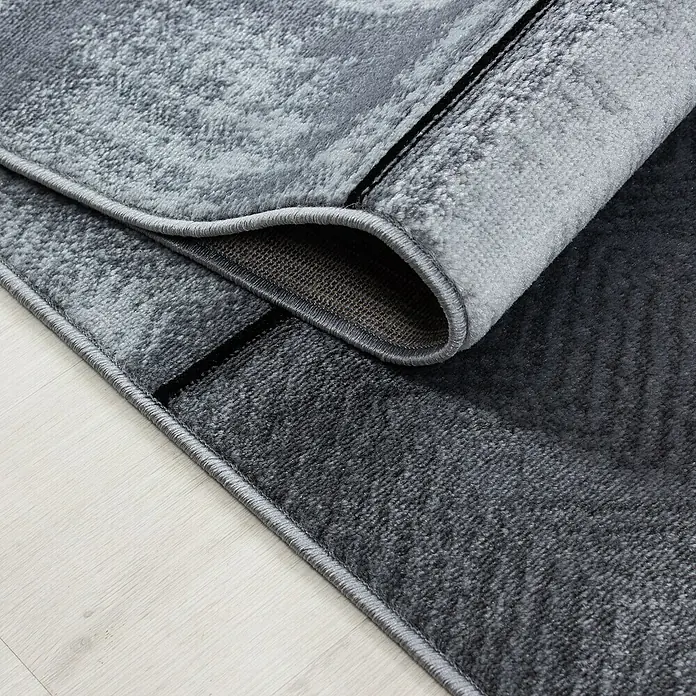 Kurzflorteppich Plus - Anthrazit, 340 x 240 cm, 100 % Polypropylen - Nahaufnahme eines modernen grauen Teppichs mit einem abstrakten Muster und einer gerollten Ecke, die seine Textur zeigt.