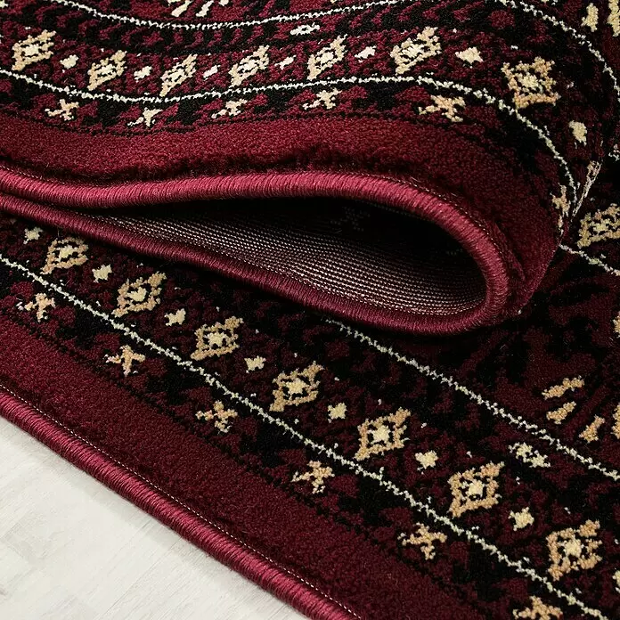 Kurzflorteppich Orient-Optik Marrakesh 0351 - Rot, 170 x 120 cm, 100% Polypropylen - Nahaufnahme eines burgunderroten Teppichs mit traditionellem Schwarz-Beige-Muster, teilweise aufgerollt.
