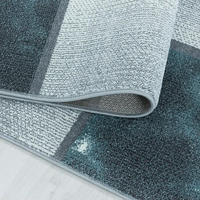 Kurzflorteppich Ottawa 4201Blau, 250 x 80 cm, 100% Polypropylen - Detailaufnahme eines Teppichs mit modernem geometrischen Muster in Grau- und Blautönen, der aufgerollt ist.