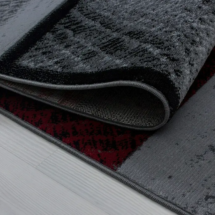 Kurzflorteppich Plus - Rot/Grau, 340 x 240 cm, 100 % Polypropylen - Nahaufnahme eines modernen grauen Teppichs mit schwarzen und roten Akzenten, teilweise aufgerollt, um die Textur zu zeigen.