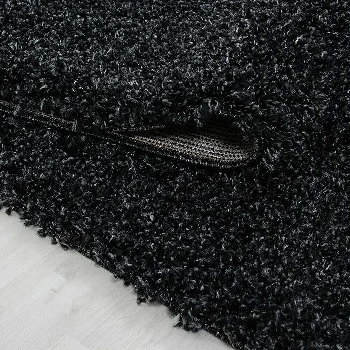 Hochflorteppich Life 1500 - Anthrazit, 110 x 60 cm, 100% Polypropylen - Nahaufnahme eines schwarzen, zotteligen Teppichs, der auf einem hellen Holzboden liegt und eine luxuriöse und komfortable Textur zeigt.