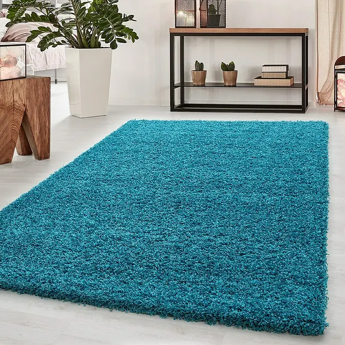 Hochflorteppich Dream 4000 - Türkis, 290 x 200 cm, 100% Polypropylen - Ein türkisfarbener, hochfloriger Teppich liegt in einem Raum mit Pflanzen und Möbeln. Der Teppich verleiht dem Raum eine gemütliche und farbenfrohe Note.