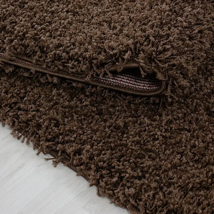 Hochflorteppich Life 1500Braun, 150 x 80 cm, 100 % Polypropylen - Nahaufnahme eines braunen, zotteligen Teppichs mit hochgezogener Ecke, der die Unterseite zeigt. Weiche Textur und warme Farbe.