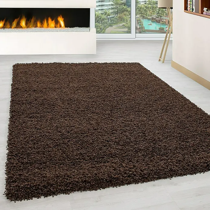 Hochflorteppich Life 1500Braun, 150 x 80 cm, 100 % Polypropylen - Ein weicher, brauner Shaggy-Teppich liegt auf einem hellen Holzboden vor einem modernen Kamin und einer Fensterfront.