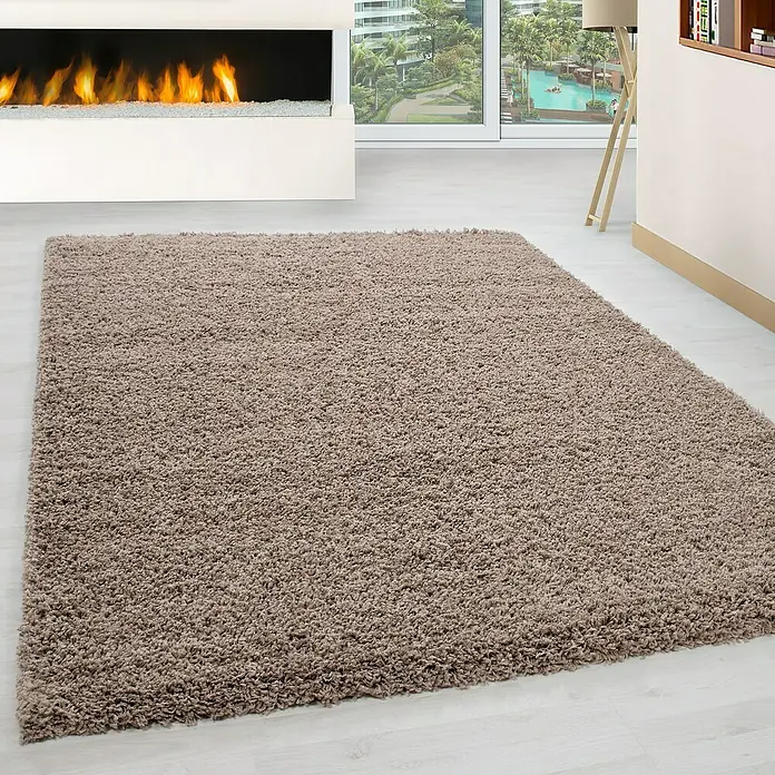 Hochflorteppich Life 1500 - Beige, 340 x 240 cm, 100% Polypropylen - Ein luxuriöser, zotteliger Teppich in einem warmen Braunton liegt in einem modernen Wohnzimmer mit Kamin und Fensterfront.