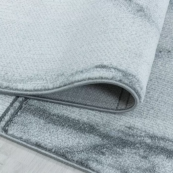 Kurzflorteppich Naxos 3816 - Silber, 200 x 140 cm, 80% Polypropylen, 20% Polyester - Heller grauer Teppich mit weicher, plüschiger Textur und modernem abstraktem Muster, teilweise aufgerollt, um Kante und gewebte Rückseite zu zeigen.
