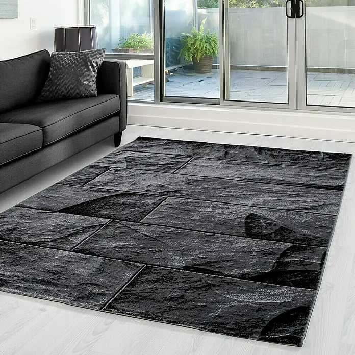 Kurzflorteppich Parma 9250 - Schwarz, 300 x 80 cm, 100% Polypropylen - Nahaufnahme eines schwarzen Teppichs mit Steinmuster, der neben einem grauen Sofa auf einem Holzboden liegt.