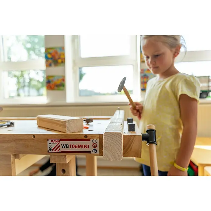 Holzmann Kinder-Werkbank WB106 Mini - L x B: 1.060 x 610 mm, Arbeitshöhe: 700 mm - Ein Mädchen benutzt einen Hammer auf einer Holzwerkbank der Marke Holz Mann WB106MINI. Die Werkbank ist aus hellem Holz gefertigt.