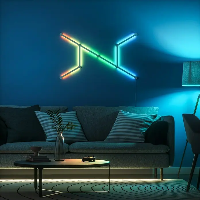 Nanoleaf Erweiterungskit Lines 60 Degrees 3 Lines - 3 Stk., Weiß, RGBW - Wandmontierte, mehrfarbige LED-Leuchte in einem Wohnzimmer. Das Licht akzentuiert die blaue Wand über einem grauen Sofa mit Kissen.