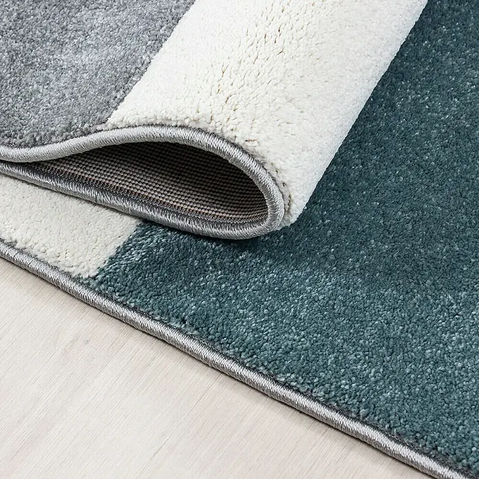 Kurzflorteppich Hawaii 1310 - Blau, 340 x 240 cm, 100 % Polypropylen - Nahaufnahme eines dekorativen Teppichs mit geometrischem Muster in Grau-, Weiß- und Blautönen, der teilweise aufgerollt ist.