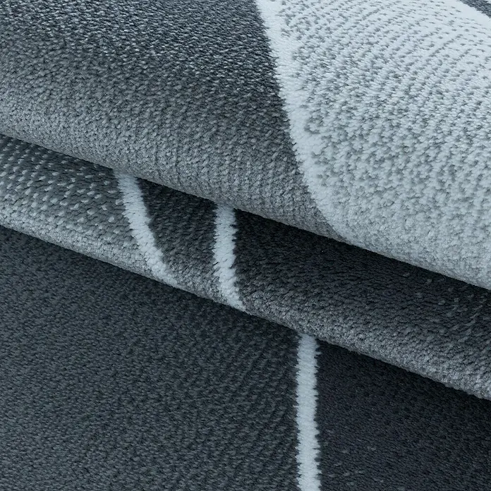 Kurzflorteppich Costa 3523Grau, 290 x 200 cm, 100 % Polypropylen - Detailaufnahme eines modernen, grau-weißen Teppichs mit geometrischem Muster, der zusammengerollt ist.